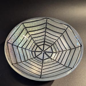 Op de afbeelding: Een heldere, ovale glazen schaal met een zwart spinnenwebontwerp. De schaal heeft een glinsterende, iriserende afwerking. Het spinnenwebontwerp is gecentreerd en bestaat uit zwarte lijnen. De schaal is waarschijnlijk voor decoratieve doeleinden.