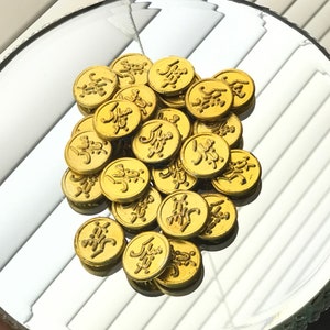 May include: A collection of small, round, gold-colored coins with embossed Arabic script. The coins are scattered on a reflective surface, creating a shimmering effect. The coins appear to be made of metal.