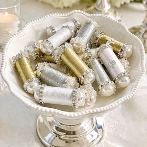 May include: A white, footed serving dish filled with decorative spools. The spools are wrapped in gold, silver, and white thread, and adorned with pearls and rhinestones. The dish sits on a silver base, creating an elegant display.