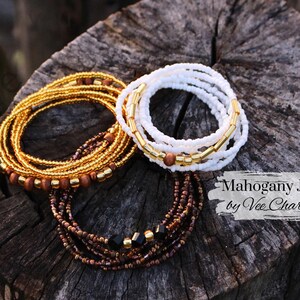 May include: Two sets of beaded waist beads, one in gold and brown with wooden beads, and one in white with gold accents. The text "Mahogany Joy by Vee Charles" is visible in the image.