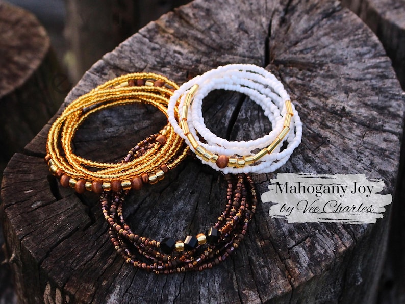 May include: Two sets of beaded waist beads, one in gold and brown with wooden beads, and one in white with gold accents. The text "Mahogany Joy by Vee Charles" is visible in the image.