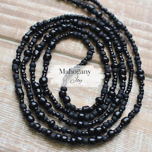 May include: A black beaded waist chain with multiple strands of beads in different sizes. The chain is on a wooden surface. The text "Mahogany Joy" is visible on a white label.