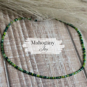 May include: A green and light green beaded necklace on a wooden surface. The necklace is labeled "Mahogany Joy".
