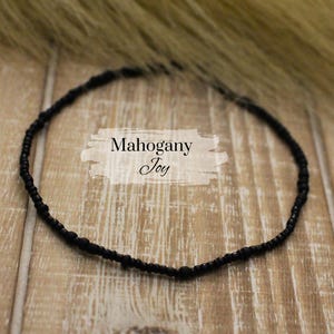 May include: A black beaded necklace with the text "Mahogany Joy" on a white background. The necklace is circular and rests on a wooden surface. The beads are small and evenly spaced.