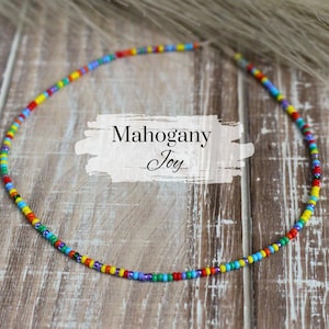 May include: A colorful beaded necklace with a variety of colors, including red, yellow, blue, green, and purple. The necklace is on a wooden surface and has the text "Mahogany Joy" written on a white background.