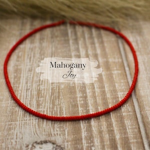 May include: A vibrant red necklace, circular in shape, rests on a light brown wooden surface. The necklace is a continuous loop of tightly woven material. The text "Mahogany Joy" is displayed in a white banner.