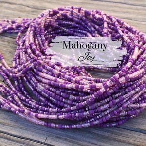May include: A collection of purple and pink beaded strands, with the text "Mahogany Joy" on a white background. The beads are a mix of sizes and shades, creating a textured and colorful display. The beads are arranged on a wooden surface.