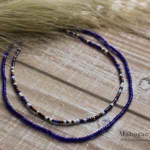 May include: Two beaded necklaces. The top necklace has white, gold, and brown beads. The bottom necklace is a solid dark blue. The necklaces are on a wooden surface with a light brown feathery accent in the background. The text "Mahogany Joy" is in the bottom right corner.