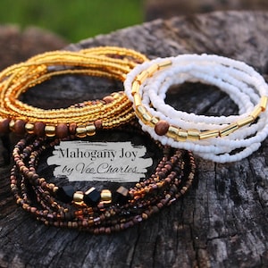 May include: Three sets of beaded bracelets in gold, white, and brown with black beads. The bracelets are stacked on top of each other and the text "Mahogany Joy by Vee Charles" is visible in the center of the image.