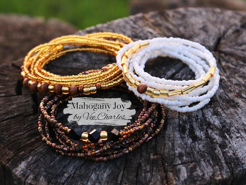 May include: Three sets of beaded bracelets in gold, white, and brown with black beads. The bracelets are stacked on top of each other and the text "Mahogany Joy by Vee Charles" is visible in the center of the image.
