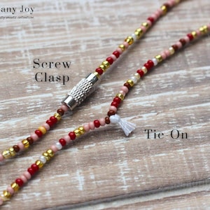May include: Close-up of a beaded necklace with a screw clasp and tie-on closure. The necklace features red, gold, pink, and white beads. The text "Mahogany Joy" and "Screw Clasp" and "Tie-On" are visible.