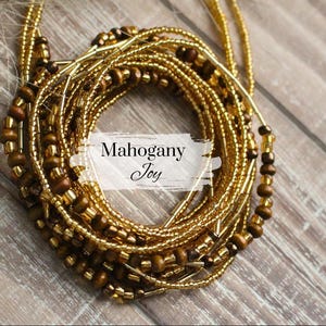 May include: A close-up of a gold and brown beaded waist chain. The chain is made of multiple strands of beads, including small gold beads and larger brown beads. The chain is arranged in a circle, and the text "Mahogany Joy" is visible in the center.