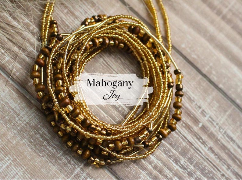 May include: A close-up of a gold and brown beaded waist chain. The chain is made of multiple strands of beads, including small gold beads and larger brown beads. The chain is arranged in a circle, and the text "Mahogany Joy" is visible in the center.