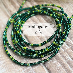 May include: A green and gold beaded waist chain, also known as a waist bead or belly chain. The chain is made of small, round beads and is arranged in a layered pattern. The text "Mahogany Joy" is visible in the image.