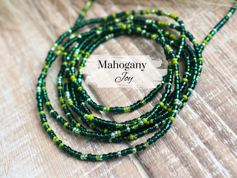 May include: A green and gold beaded waist chain, also known as a waist bead or belly chain. The chain is made of small, round beads and is arranged in a layered pattern. The text "Mahogany Joy" is visible in the image.