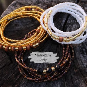 May include: Two sets of beaded waist beads, one in gold and brown, and the other in white and gold. The gold and brown set has a tag that reads "Mahogany Joy".
