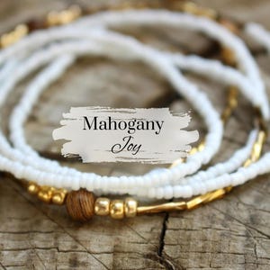 May include: White beaded necklace with gold accents and a brown wooden bead. The necklace is layered and has a "Mahogany Joy" label.