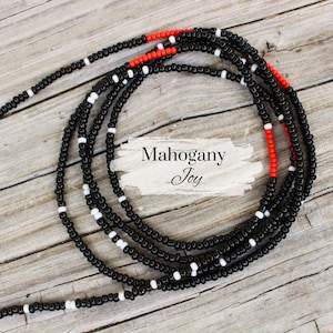 May include: Strands of black waist beads with white and red accents. The beads are arranged in a circular pattern on a wooden surface. The text "Mahogany Joy" is displayed on a white background.