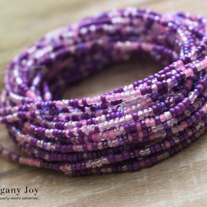 May include: A coiled strand of waist beads in shades of purple, pink, and clear. The beads are small and uniform in size, creating a textured appearance. The beads are likely strung on a durable cord. The words "Mahogany Joy" are at the bottom of the image.