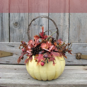 May include: A white pumpkin with a wire handle is filled with a fall floral arrangement. The arrangement includes brown pine cones, red and orange leaves, and pink flowers. The pumpkin is sitting on a weathered wooden surface.