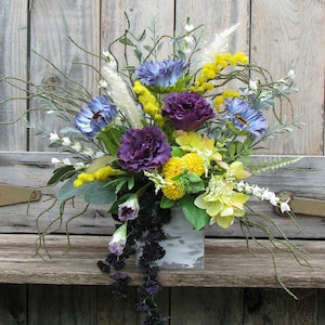 May include: A floral arrangement in a whitewashed, rustic planter. The arrangement features purple carnations, blue and yellow flowers, and sprigs of greenery.