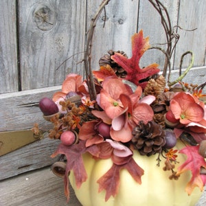 May include: A white pumpkin with a brown vine handle is decorated with fall foliage, including red, pink, and brown leaves, pinecones, and acorns.