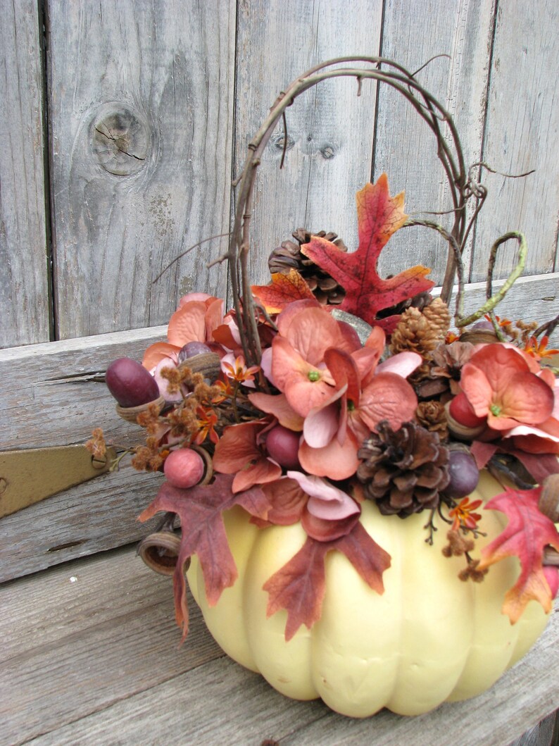 May include: A white pumpkin with a brown vine handle is decorated with fall foliage, including red, pink, and brown leaves, pinecones, and acorns.