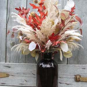 May include: A decorative fall arrangement in a dark brown glass bottle. The arrangement features red, orange, and cream-colored dried leaves and grasses. The bottle is cylindrical with a wide base and a narrow neck.