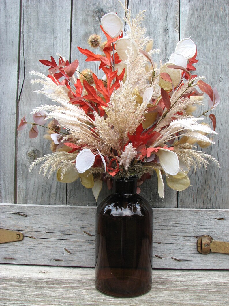 May include: A decorative fall arrangement in a dark brown glass bottle. The arrangement features red, orange, and cream-colored dried leaves and grasses. The bottle is cylindrical with a wide base and a narrow neck.
