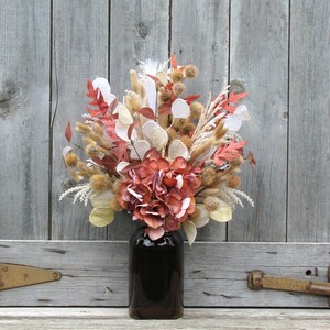 May include: A bouquet of dried flowers in a dark brown glass bottle. The arrangement includes red, white, and tan elements, with a large, reddish-brown hydrangea as the focal point. The background is a weathered, gray wooden surface.