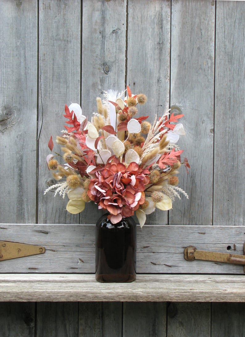 May include: A bouquet of dried flowers in a dark brown glass bottle. The arrangement includes red, white, and tan elements, with a large, reddish-brown hydrangea as the focal point. The background is a weathered, gray wooden surface.