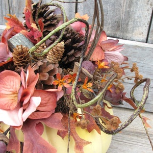May include: A yellow pumpkin with a fall-themed arrangement of dried leaves, pine cones, and flowers. The arrangement includes shades of brown, orange, and pink.