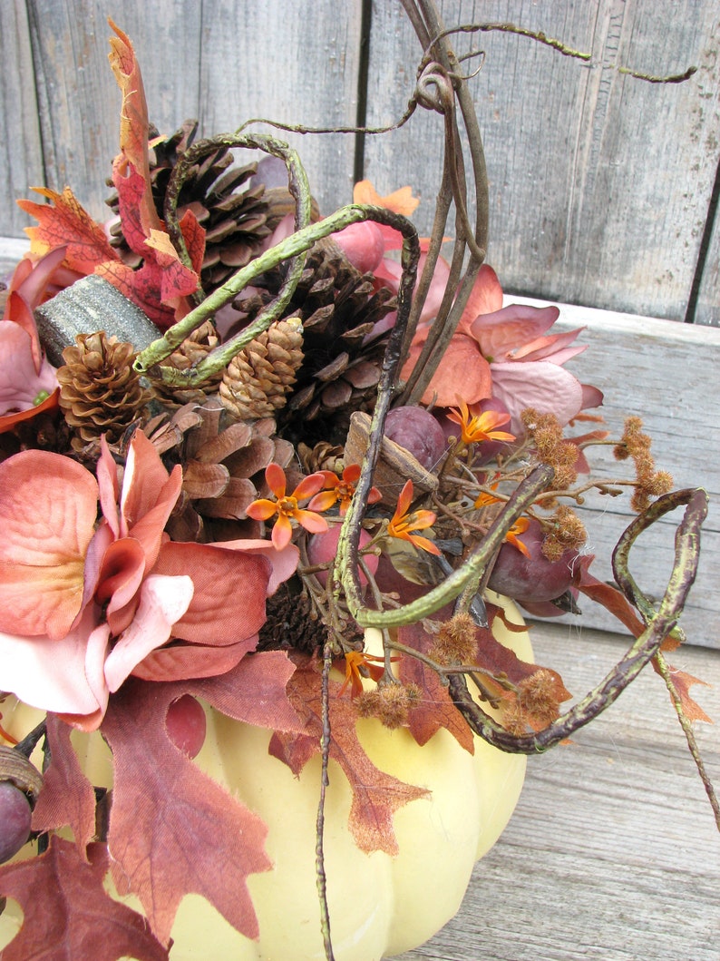 May include: A yellow pumpkin with a fall-themed arrangement of dried leaves, pine cones, and flowers. The arrangement includes shades of brown, orange, and pink.
