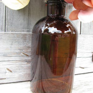 May include: A large, brown glass bottle with a rounded body and a narrow neck. The bottle has a glossy finish, reflecting the surrounding environment. The bottle is likely used for decorative purposes or as a vase. The bottle is sitting on a wooden surface.