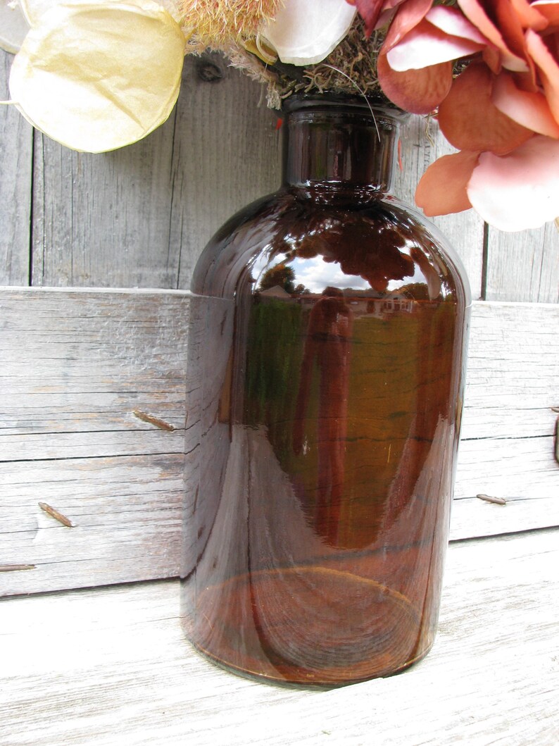 May include: A large, brown glass bottle with a rounded body and a narrow neck. The bottle has a glossy finish, reflecting the surrounding environment. The bottle is likely used for decorative purposes or as a vase. The bottle is sitting on a wooden surface.