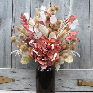 May include: A fall-themed floral arrangement in a dark brown glass bottle. The bouquet features dried flowers and foliage in shades of orange, brown, cream, and white. The arrangement is set against a weathered wooden background.