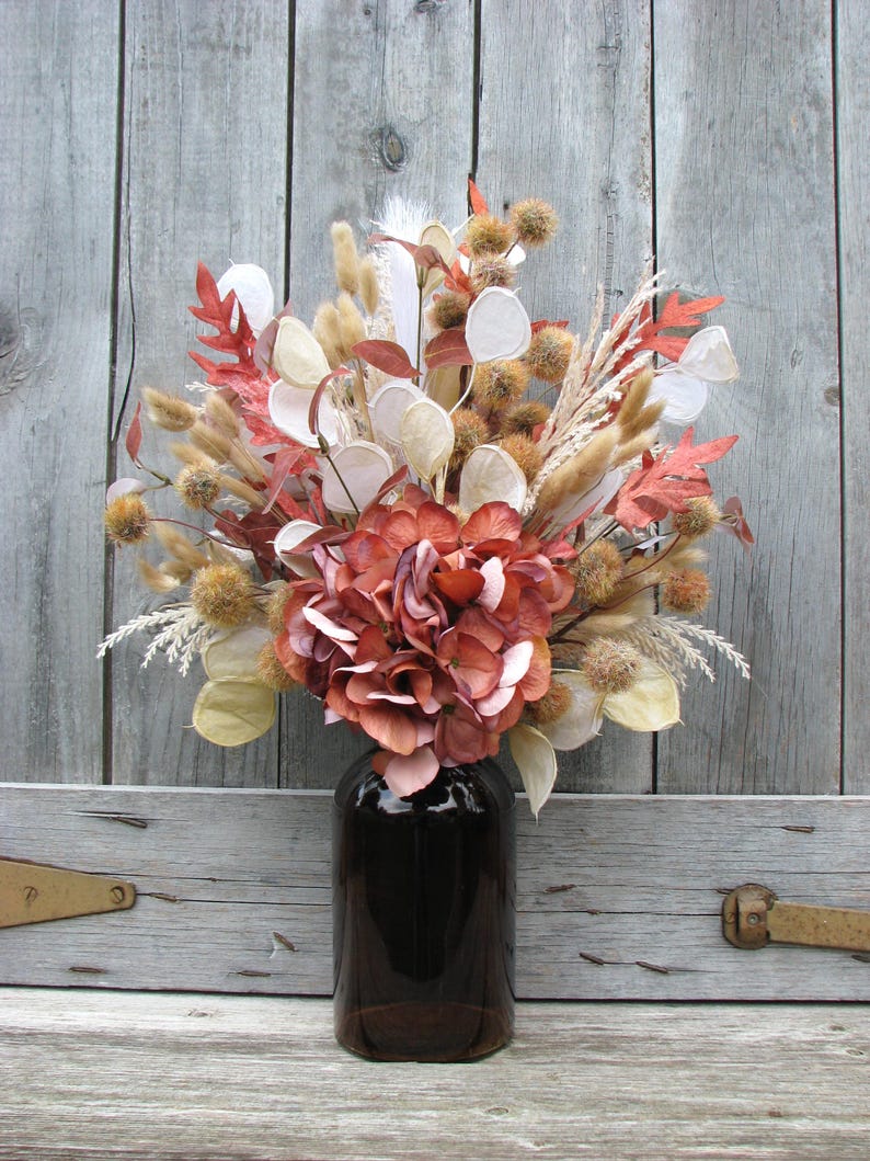 May include: A fall-themed floral arrangement in a dark brown glass bottle. The bouquet features dried flowers and foliage in shades of orange, brown, cream, and white. The arrangement is set against a weathered wooden background.