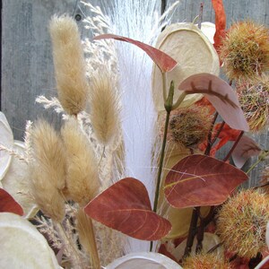 May include: A close-up of a dried floral arrangement featuring various textures and colors. The arrangement includes tan bunny tails, white feathery plumes, and reddish-brown leaves. Other elements include seed pods and spiky, orange-toned accents.