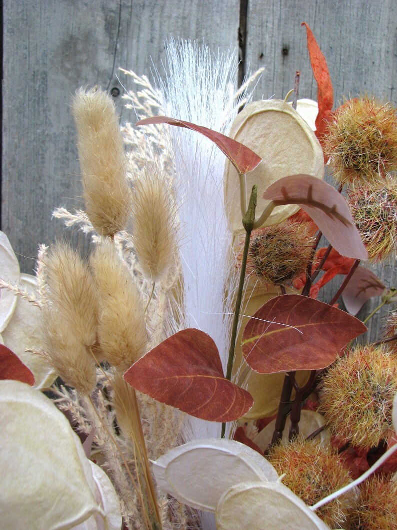 May include: A close-up of a dried floral arrangement featuring various textures and colors. The arrangement includes tan bunny tails, white feathery plumes, and reddish-brown leaves. Other elements include seed pods and spiky, orange-toned accents.