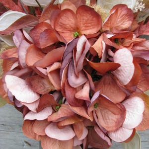 May include: A close-up of a bouquet of artificial flowers in shades of peach, mauve, and white. The flowers have a layered petal design and are arranged in a dark brown vase. The overall theme is floral and decorative.