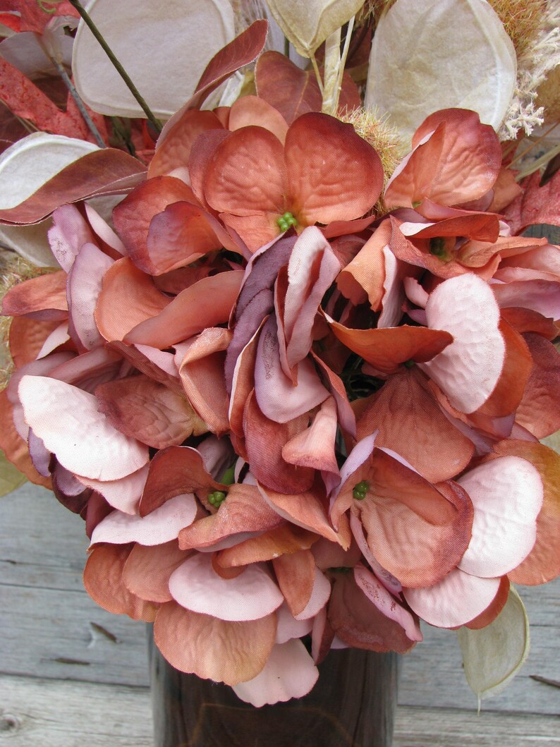 May include: A close-up of a bouquet of artificial flowers in shades of peach, mauve, and white. The flowers have a layered petal design and are arranged in a dark brown vase. The overall theme is floral and decorative.
