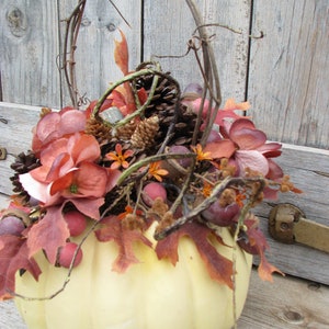 May include: A white pumpkin with a vine wrapped around it. The pumpkin is filled with fall foliage, pine cones, and berries.