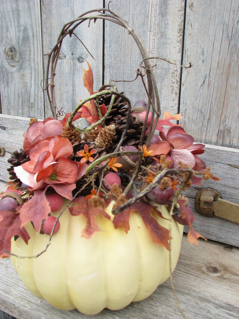 May include: A white pumpkin with a vine wrapped around it. The pumpkin is filled with fall foliage, pine cones, and berries.