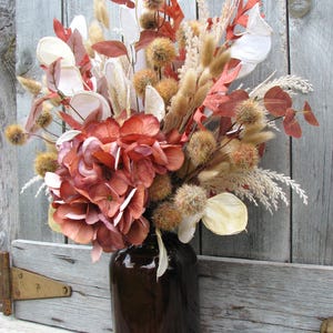 May include: A brown glass vase filled with a dried floral arrangement. The arrangement includes large, rust-colored hydrangea blooms, tan grasses, and various dried seed pods and leaves in shades of brown, cream, and orange. The vase sits against a weathered wooden background.