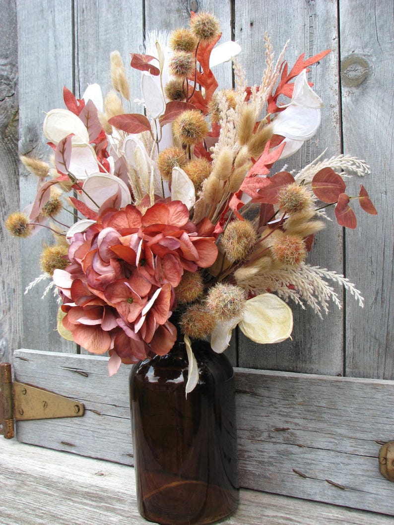 May include: A brown glass vase filled with a dried floral arrangement. The arrangement includes large, rust-colored hydrangea blooms, tan grasses, and various dried seed pods and leaves in shades of brown, cream, and orange. The vase sits against a weathered wooden background.