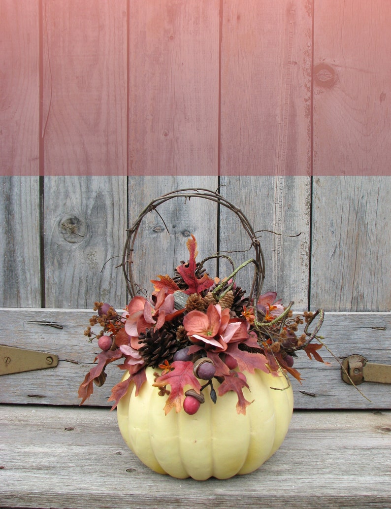 May include: A white pumpkin with a wire handle is filled with a fall floral arrangement. The arrangement includes brown pine cones, red and orange leaves, and pink flowers. The pumpkin is sitting on a weathered wooden surface.