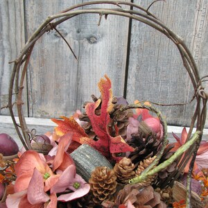 May include: A fall-themed arrangement in a twig wreath with pine cones, faux leaves, and pink flowers.