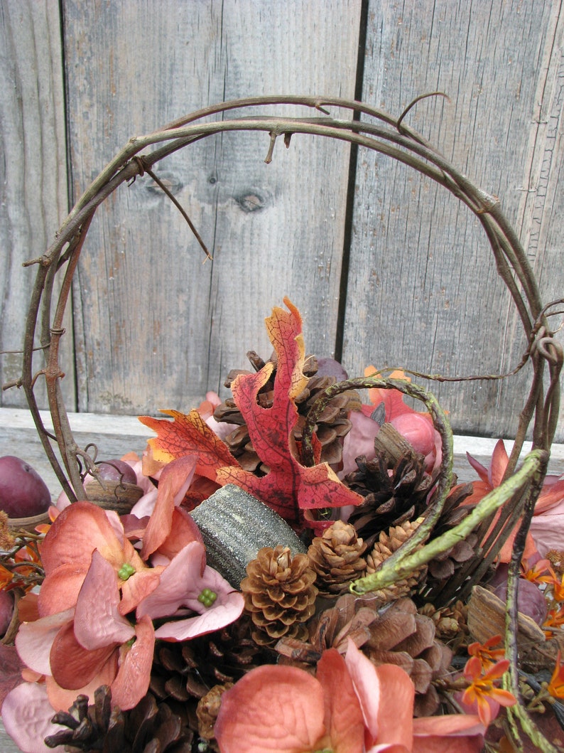 May include: A fall-themed arrangement in a twig wreath with pine cones, faux leaves, and pink flowers.