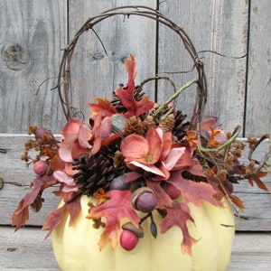 May include: A pale yellow pumpkin with a wire handle is decorated with fall foliage, pinecones, and berries. The pumpkin is sitting on a wooden surface.