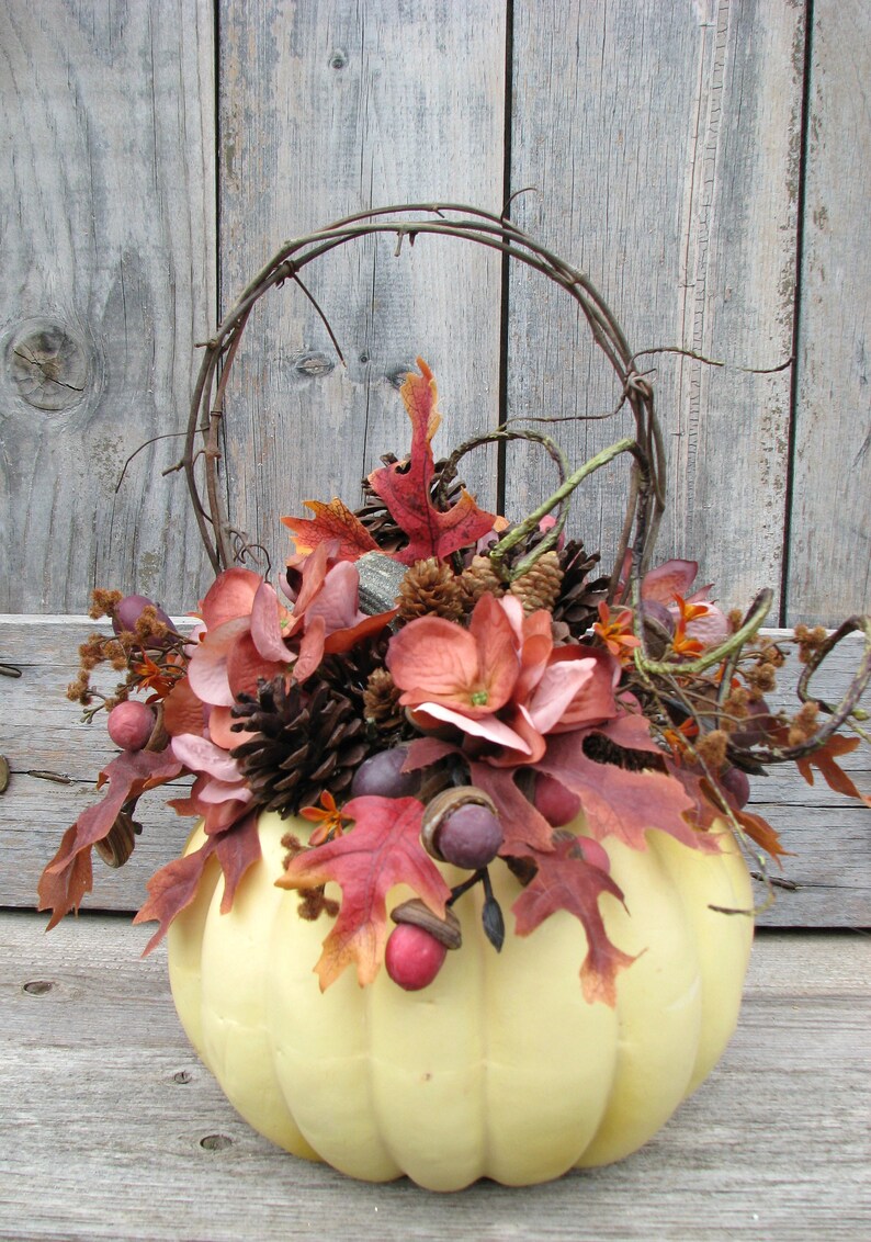 May include: A pale yellow pumpkin with a wire handle is decorated with fall foliage, pinecones, and berries. The pumpkin is sitting on a wooden surface.